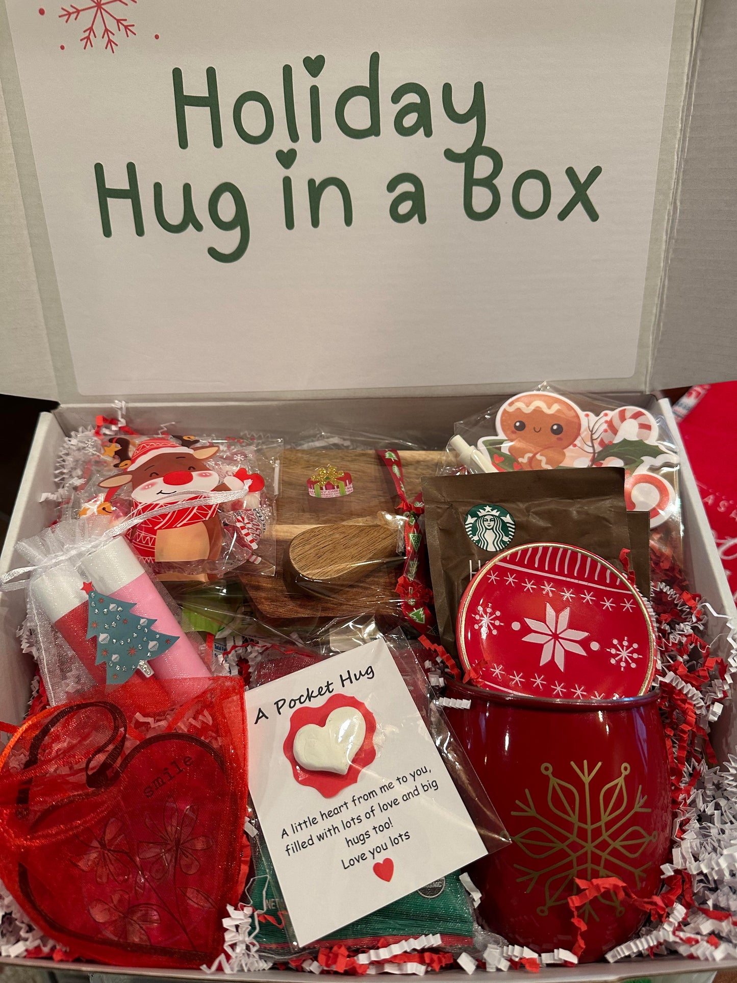 Box labeled 'Holiday Hug in a Box' with Starbucks mug, heart-shaped pouch, and other items.